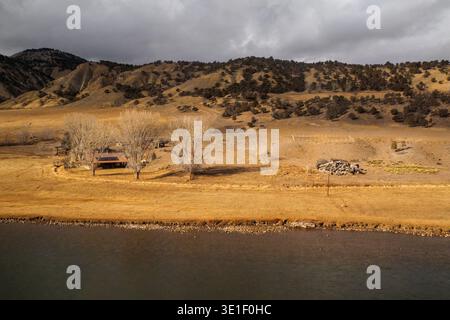 A remote ranch house sits nestled in a dry, golden valley, with barren trees and scrub brush dotting the landscape. The scene is set against a backdrop of rugged, eroded hills under a dramatic, cloudy sky, with a dark body of water in the foreground. Stock Photo