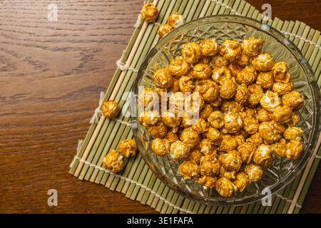 A bowl holds popcorn placed on a wooden table. The popcorn looks shiny and is surrounded by a bamboo mat. The setting shows a simple snack presentatio Stock Photo