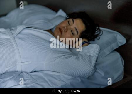 A young Hispanic woman with dark hair relaxes in bed, wearing a white robe. She appears peaceful, with her eyes closed and hands behind her head. Stock Photo