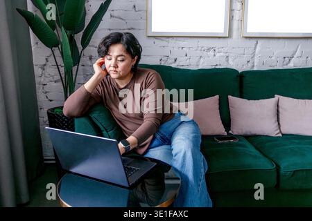 A young Hispanic woman with curly black hair sits on a green couch, working on a laptop. She wears a brown sweater and blue jeans, looking focused and Stock Photo