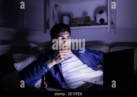 Frightened man eating pizza and watching movie on laptop Stock Photo