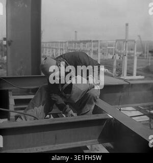 Vintage 1960s photograph capturing a highly focused Soviet construction worker welding a large steel beam at an expanding industrial building site in the Slavyansk region. The skilled assembler, looking carefully through his lowered protective mask, uses an electrode holder to precisely fuse the heavy metal framework. An authentic historical archive image highlighting the technical precision, harsh manual labor, and massive engineering scale. A poignant glimpse into life before the war and peaceful Donbas. Stock Photo