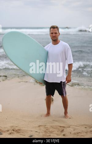 Full Body Shot Of Man At Seaside. Relaxed Individual In White Shirt With Surfboard On Sandy Coast Stock Photo