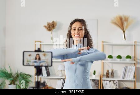 Smiling young woman recording fitness video on smartphone and showing warm up exercise at home. Stock Photo