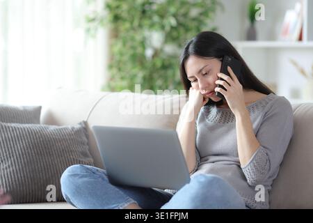 Bored asian woman calling on phone checking laptop sitting on a sofa at home Stock Photo