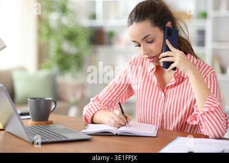 Freelance worker talking on phone writing in paper agenda working at home Stock Photo