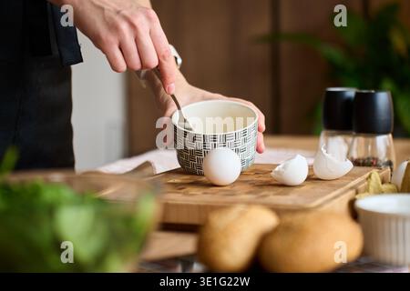 Hand mixing beaten eggs in ceramic bowl on kitchen counter with eggshells and wooden cutting board. Person preparing healthy breakfast Stock Photo