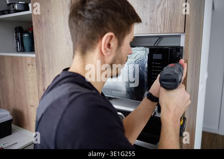 Man using cordless screwdriver to fix built in new microwave oven in modern kitchen cabinet. Concept of home appliance using Stock Photo