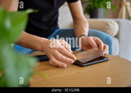 Man holding smartphone and applying protective glass screen protector while sitting by wooden table at home. Concept of device protection, mobile acce Stock Photo