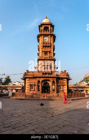 The ornate Ghanta Ghar clock tower stands centrally in a paved square of Jodhpur as a woman in red attire walks past the base. Stock Photo