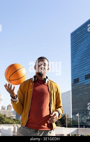 African American man standing in city plaza spinning basketball with headphones and yellow jacket Stock Photo