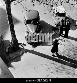 Captured by Max Dupain in the 1930s or 1940s, this black and white photograph from Adelaide presents an elevated view of a street scene. A vintage car is parked beside a tree, while two men converse near another car and a third individual walks away, creating dynamic shadows on the paved ground. Stock Photo