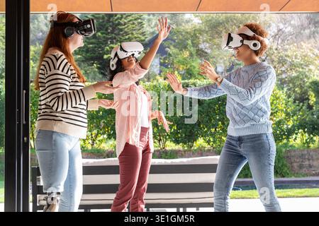 Diverse female friends interacting with virtual objects near bench on patio using VR headsets. Technology, innovation, collaboration, outdoor, leisure, modern, connectivity || Model released Stock Photo