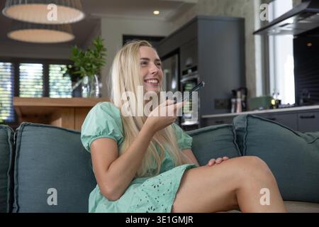Woman sitting on teal sofa in modern open living area holding remote control and observing greenery. Contemporary, minimalist, relaxing, interior, lifestyle, home decor, vibrant || Model released Stock Photo