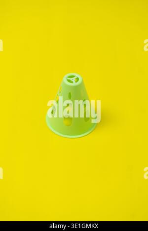 A single light green perforated plastic cone stands centrally on a vibrant yellow surface, creating a striking and minimalist composition. Stock Photo