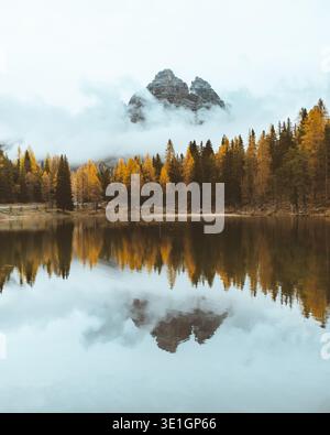 View of golden trees reflecting on the calm lake, under a sky where clouds dance around a majestic mountain peak, creating a serene landscape, Misurin Stock Photo