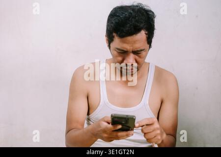 A man in a white tank top is intently focused on his smartphone. His serious and concentrated expression suggests he is reading important messages or Stock Photo