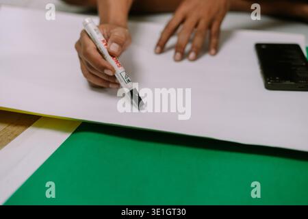 A close-up shot of a person's hand holding a black marker, poised over a large blank white sheet of paper with a green background. This image conveys Stock Photo