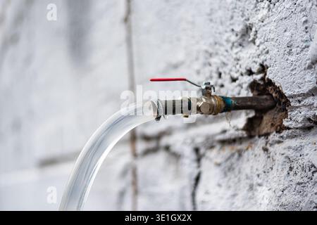 A metal tap sticks out of the white rough wall with water pouring in a transparent hose. High quality photo Stock Photo