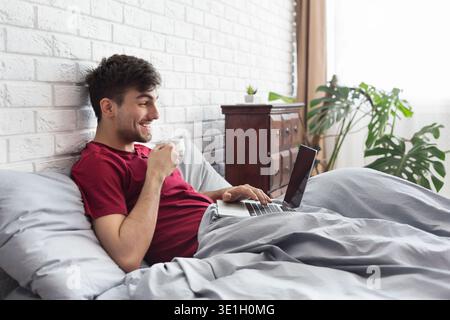 Happy man drinking coffee and using laptop in bed Stock Photo
