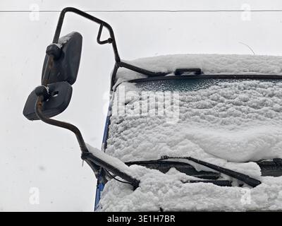 Snow covers the top of a vehicle, including the windshield and side mirror. The scene shows a cold winter day with heavy snowfall on the surface. Stock Photo