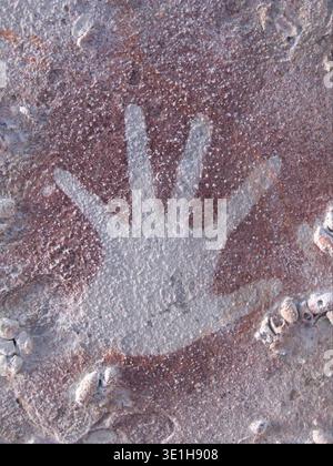 This ancient rock art, likely dating back thousands of years to the prehistoric era, features a striking white hand stencil against a reddish-brown cave wall. The technique involves blowing pigment around an outstretched hand to create a negative impression, a widespread form of early human artistic expression. Stock Photo