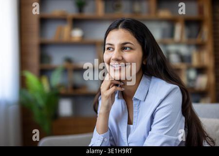 Young indian woman sitting on a sofa in her modern living room, smiling and looking away, pondering new ideas or enjoying a peaceful moment of relaxation and contemplation Stock Photo