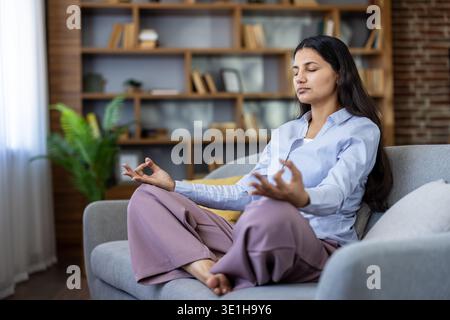 Young woman sitting barefoot in lotus pose on a cozy couch at home, practicing meditation and mindfulness for calm, inner peace, stress relief and overall wellbeing Stock Photo