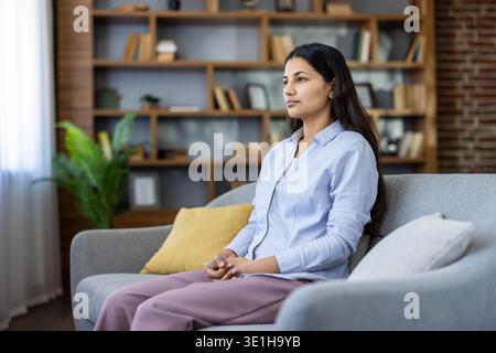 Young woman sitting on a comfortable sofa in a modern living room, looking away thoughtfully while contemplating or reflecting on future plans and strategies Stock Photo