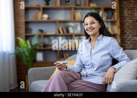 Happy woman sitting on sofa and smiling, feeling overjoyed and excited finding out positive pregnancy test result, embracing future motherhood and new beginnings Stock Photo