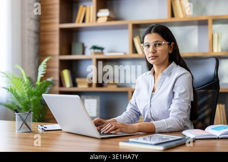 Young professional businesswoman wearing glasses sitting at a wooden desk, working on a laptop while looking at the camera, with a modern bookshelf and plant in the blurred background Stock Photo
