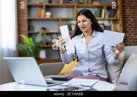 Young woman at home stressed over financial bills and receipts, sitting on sofa with laptop and paperwork, worried about debt, budgeting and expenses while working remotely Stock Photo