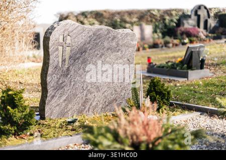 A grave marker stands in a cemetery with green trees and flower arrangements nearby. The sun shines on the stones, highlighting the surroundings. The Stock Photo