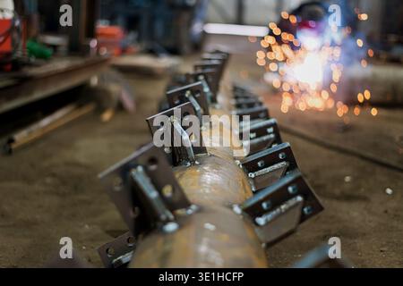 A large pipe with welded blades lies on the floor of the workshop. In the background, a welder is welding metal by scattering sparks. High quality pho Stock Photo
