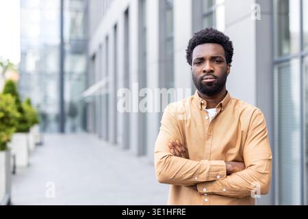 Young business man standing with a thoughtful and determined expression, crossing his arms in a powerful stance outside a modern corporate building, representing ambition and success Stock Photo
