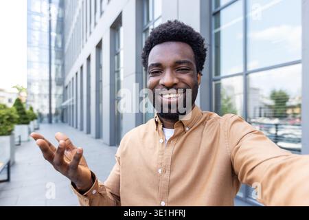 Young adult african american man with a beard smiling happily and gesturing with his hand while recording a video call, or taking a selfie in front of a contemporary office exterior Stock Photo