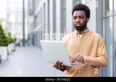 Young african man standing outside a modern office building, focused on his laptop and typing, illustrating remote work, urban connectivity, freelance tech career and professional concentration Stock Photo