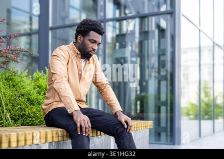 Young man sitting on an urban bench, pensive and solitary, lost in worry and fatigue amid modern glass buildings, reflecting on job loss, stress and uncertainty Stock Photo