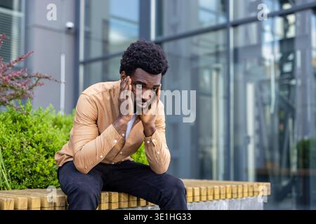 Young man feeling overwhelmed and unhappy, sitting alone outside with hands pressed to his head, indicating stress, disappointment, or mental struggle Stock Photo