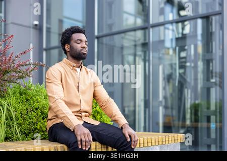 Young man with eyes closed practicing mindfulness and meditation on a bench in an urban setting, finding calm amidst city life and focusing on wellness and mental well-being Stock Photo
