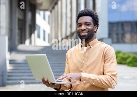 Smiling man standing outdoors holding a laptop, interacting with the device, and connecting with technology while embracing flexible work and education concepts Stock Photo