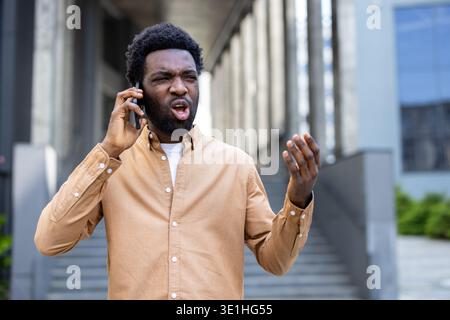 African man on a city street reacts angrily during a phone call, shouting into his mobile with visible frustration, hand gesture and tense facial expression conveying conflict and stress Stock Photo