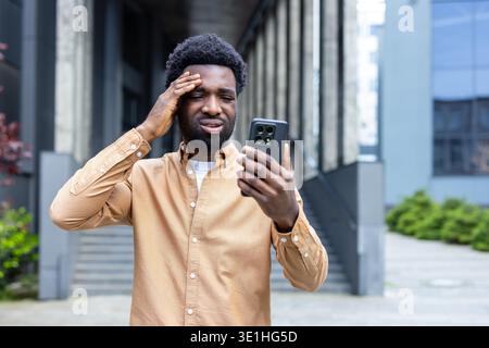 Stressed young person clenching head while looking at a cellphone with a worried expression, reacting to financial problems, business crisis, or upsetting online message Stock Photo