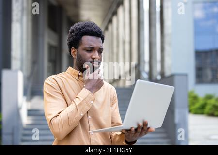 Young thoughtful male professional analyzing information on a portable computer, contemplating business strategies or solutions while standing outside a modern building Stock Photo