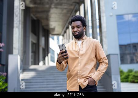 Young adult african man standing outdoors in an urban environment, browsing content on his mobile phone while connecting with technology and enjoying modern city living Stock Photo