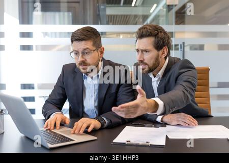 Two stressed businessmen working together, analyzing data on a laptop screen while discussing a problem, reflecting frustration and teamwork in a mode Stock Photo