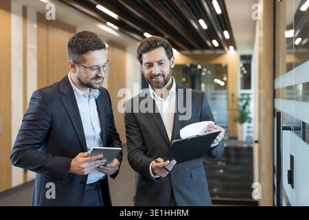 Two professional businessmen are collaborating in an office corridor, reviewing documents on a clipboard and tablet, fostering teamwork and corporate Stock Photo