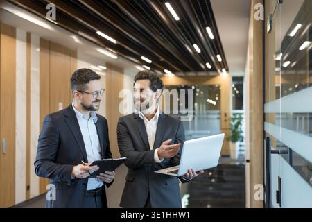 Two smiling businessmen walk through a modern corporate corridor discussing data on a laptop and clipboard, collaborating on strategy and planning in Stock Photo