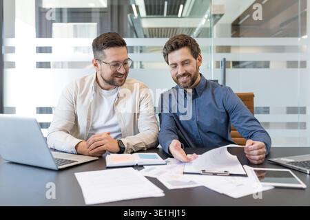 Two smiling business professionals sitting at a desk, reviewing documents and discussing project details during a productive meeting in a bright corpo Stock Photo