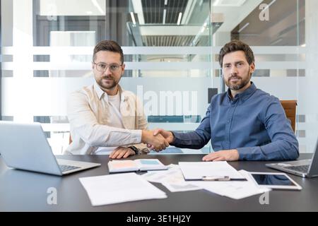 Two professional businessmen smiling, sitting at a conference table, shaking hands and concluding a successful partnership agreement in a modern offic Stock Photo